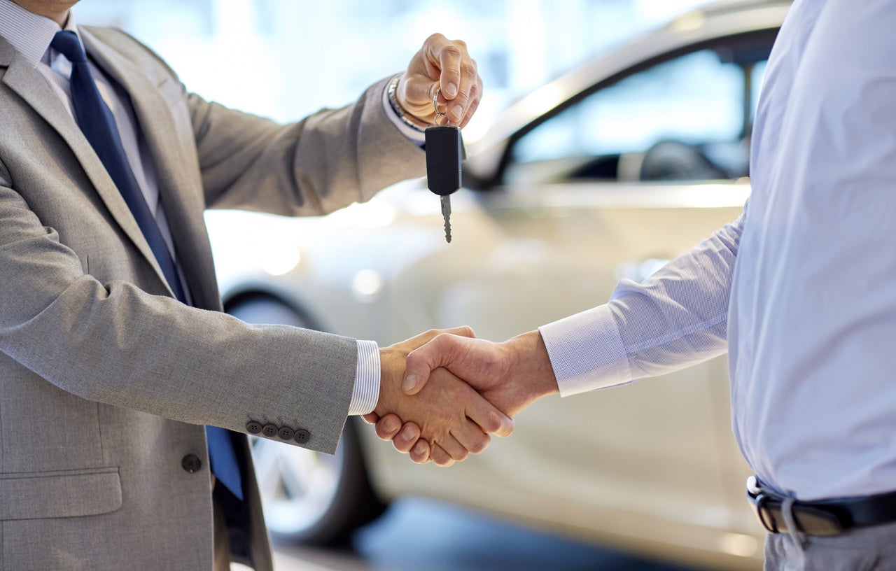 Two men shaking hands, one in a suit holding car keys, in front of a white car.