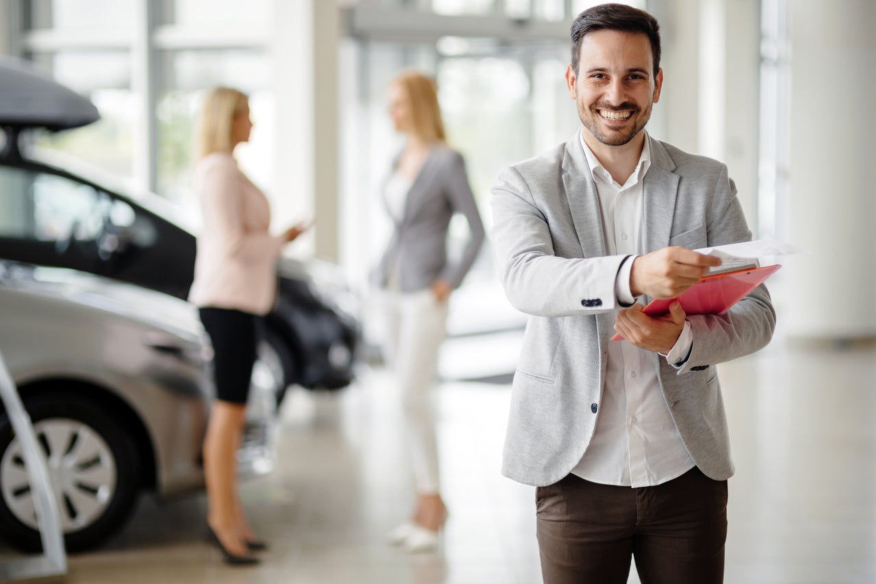 Smiling salesman in a car dealership, offering a document. Two female customers and cars are in the background.