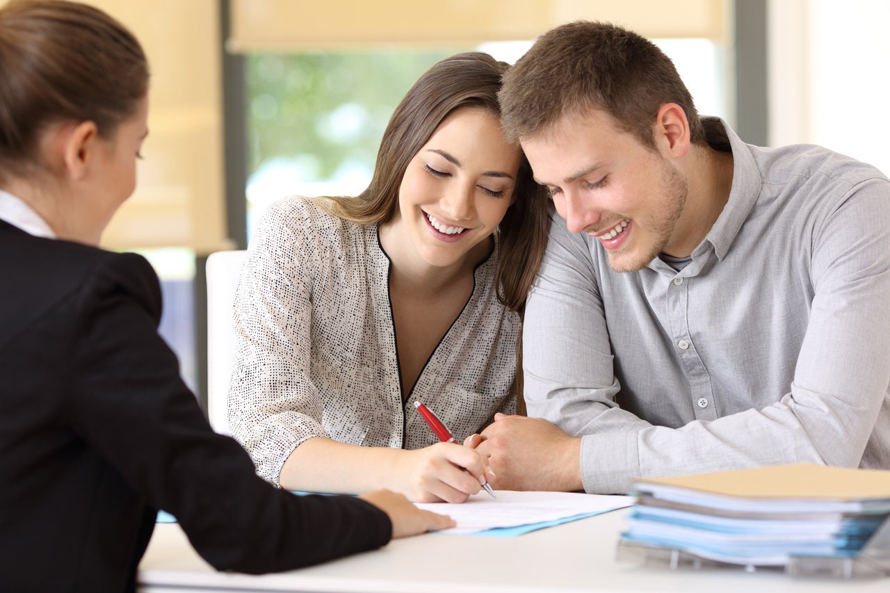 A smiling couple signs documents with a female consultant at a table, viewed from over the consultant's shoulder.