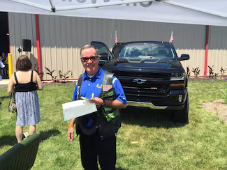 Man in blue shirt and black vest holding papers, standing in front of a black pickup truck on grass.