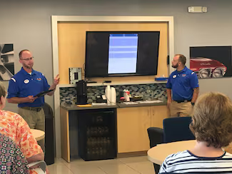 Two men in uniform present to an audience, with a monitor displaying an app's interface in the background.