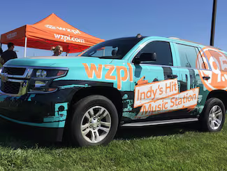 WZPL radio station SUV, turquoise and black with Indy's Hit Music Station on the side, parked on grass.