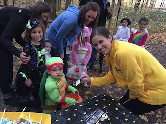 Adult handing a treat to a small child in a turtle costume, surrounded by other children in Halloween costumes.