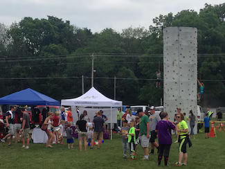 People gathered in a field for an event with tents and a tall rock climbing wall where two people are climbing.