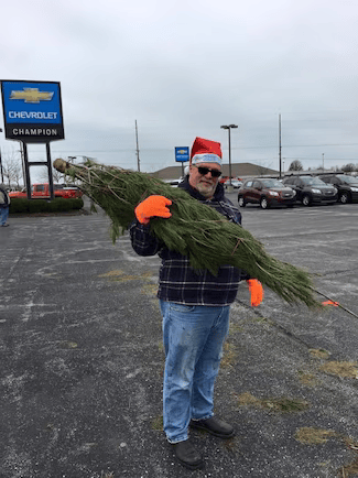 Man in Santa hat and neon orange gloves carries a bundled Christmas tree in a Chevrolet dealership parking lot.